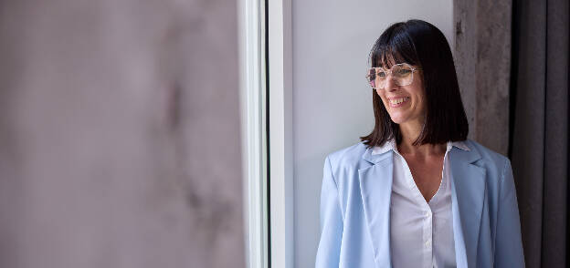 Businessportrait Frau mit hellblauem Blazer und weißer Bluse, schwarzen Haaren und Brille lächelnd am Fenster - Businessfotograf Businessfotografie Christian Vorhofer